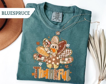 May include: Teal-colored t-shirt with a turkey graphic and the word "Thankful" in orange and brown. The turkey has patterned feathers and a polka-dot body. A gold necklace is draped over the shirt.