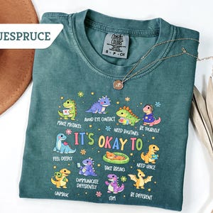 May include: A teal Comfort Colors t-shirt with a dinosaur graphic and the text "It's Okay To" with various phrases. A gold necklace and a brown hat are also visible.