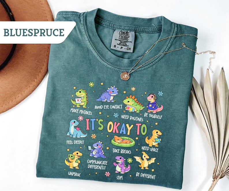 May include: A teal Comfort Colors t-shirt with a dinosaur graphic and the text "It's Okay To" with various phrases. A gold necklace and a brown hat are also visible.