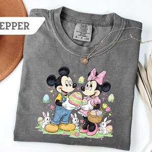 May include: Pepper-colored t-shirt featuring Mickey and Minnie Mouse holding Easter eggs. The graphic includes bunnies, flowers, and decorated eggs. The shirt has a crew neck and short sleeves. A gold necklace is draped across the shirt.