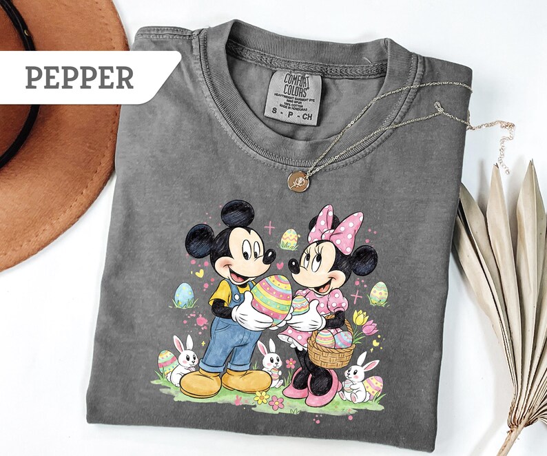 May include: Pepper-colored t-shirt featuring Mickey and Minnie Mouse holding Easter eggs. The graphic includes bunnies, flowers, and decorated eggs. The shirt has a crew neck and short sleeves. A gold necklace is draped across the shirt.