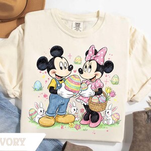 May include: Ivory-colored t-shirt featuring Mickey and Minnie Mouse holding Easter eggs. Mickey wears blue overalls and Minnie a pink polka-dot dress. Easter eggs and bunnies are scattered around the characters. The word "IVORY" is printed at the bottom.