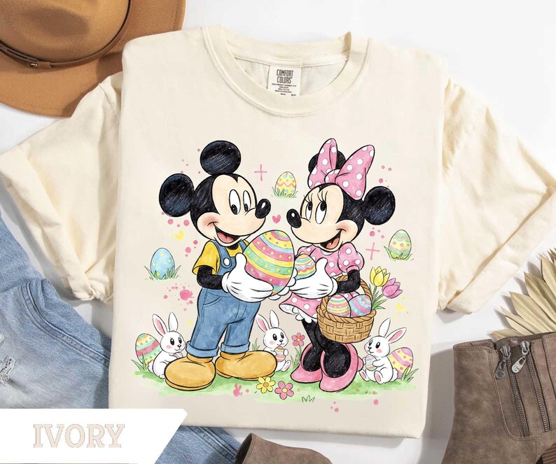 May include: Ivory-colored t-shirt featuring Mickey and Minnie Mouse holding Easter eggs. Mickey wears blue overalls and Minnie a pink polka-dot dress. Easter eggs and bunnies are scattered around the characters. The word "IVORY" is printed at the bottom.
