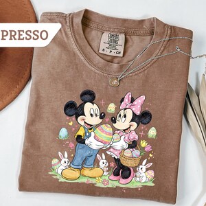 May include: Espresso-colored t-shirt featuring Mickey and Minnie Mouse holding Easter eggs. The graphic includes bunnies, flowers, and decorated eggs. A silver necklace with a pendant is draped across the shirt.