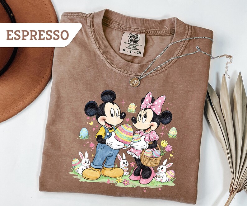 May include: Espresso-colored t-shirt featuring Mickey and Minnie Mouse holding Easter eggs. The graphic includes bunnies, flowers, and decorated eggs. A silver necklace with a pendant is draped across the shirt.
