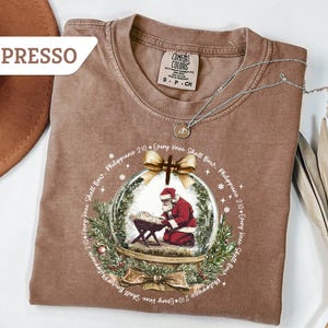 May include: Espresso-colored t-shirt with a graphic of Santa Claus kneeling in a snow globe. The design includes text from Philippians 2:10 and decorative elements. The shirt has a crew neck and short sleeves.