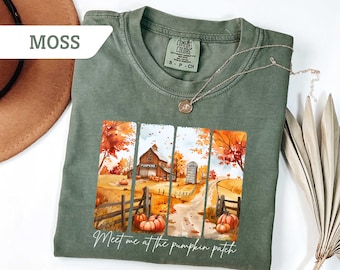 May include: Moss green t-shirt with a fall-themed graphic. The graphic features a barn, pumpkins, and the text "Meet me at the pumpkin patch." A silver necklace rests on the shirt. The shirt is made of a soft material.