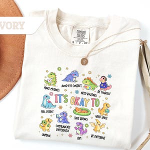 May include: Ivory-colored t-shirt with a crew neck featuring colorful cartoon dinosaurs. The shirt has the text "IT'S OKAY TO" with various phrases like "Make Mistakes" and "Be Yourself". A Comfort Colors tag is visible.
