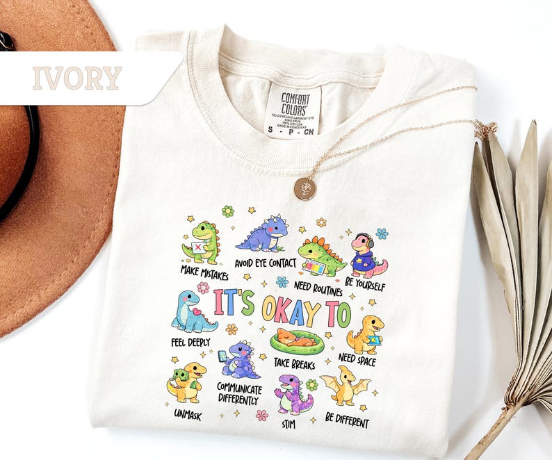 May include: Ivory-colored t-shirt with a crew neck featuring colorful cartoon dinosaurs. The shirt has the text "IT'S OKAY TO" with various phrases like "Make Mistakes" and "Be Yourself". A Comfort Colors tag is visible.
