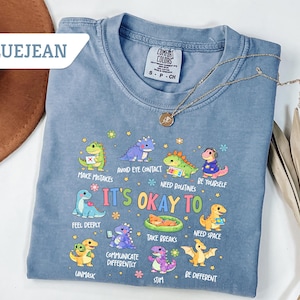 May include: Bluejean Comfort Colors t-shirt with a graphic of colorful cartoon dinosaurs and the text "It's Okay To" followed by various affirmations. A gold necklace and a brown hat are also visible.