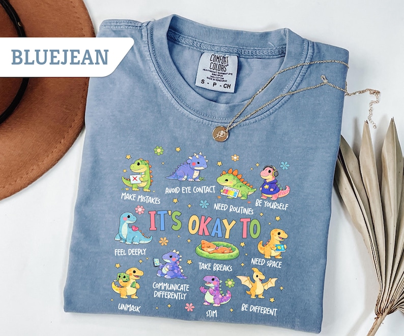 May include: Bluejean Comfort Colors t-shirt with a graphic of colorful cartoon dinosaurs and the text "It's Okay To" followed by various affirmations. A gold necklace and a brown hat are also visible.