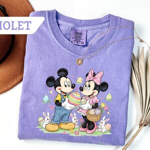 May include: Violet Comfort Colors t-shirt featuring Mickey and Minnie Mouse holding a decorated Easter egg. The graphic includes Easter eggs, bunnies, and flowers. A gold necklace is draped on the shirt.