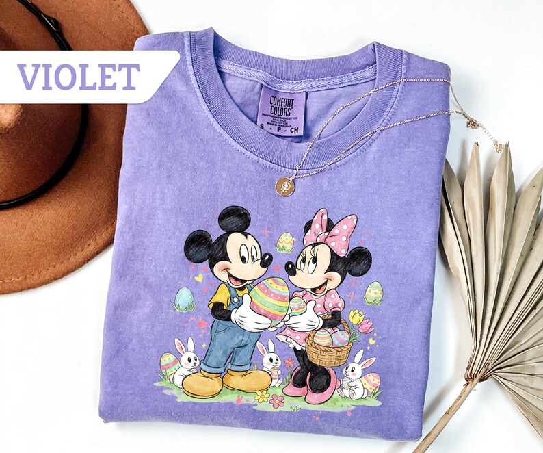 May include: Violet Comfort Colors t-shirt featuring Mickey and Minnie Mouse holding a decorated Easter egg. The graphic includes Easter eggs, bunnies, and flowers. A gold necklace is draped on the shirt.