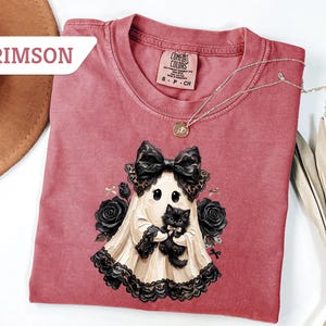 May include: A crimson t-shirt featuring a graphic of a ghost wearing a large black bow and lace trim, holding a small black kitten. The ghost is surrounded by black roses. The shirt has a Comfort Colors label and the text CRIMSON in the corner.