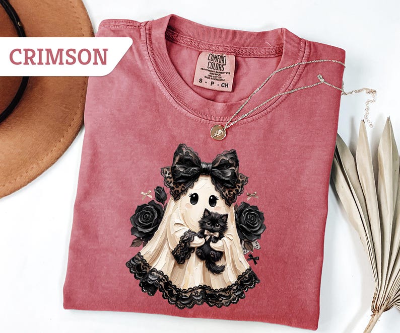 May include: A crimson t-shirt featuring a graphic of a ghost wearing a large black bow and lace trim, holding a small black kitten. The ghost is surrounded by black roses. The shirt has a Comfort Colors label and the text CRIMSON in the corner.