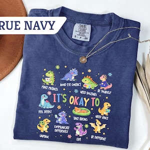 May include: A navy blue t-shirt with the text "TRUE NAVY" and a graphic of cartoon dinosaurs with the phrase "IT'S OKAY TO" and various affirmations. A gold necklace and a brown hat are also visible.
