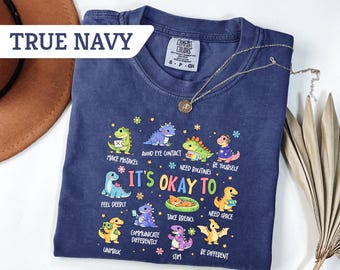May include: A navy blue t-shirt featuring the text "TRUE NAVY" and a graphic of cartoon dinosaurs with the phrase "IT'S OKAY TO" and various affirmations. A gold necklace and a brown hat are also visible.
