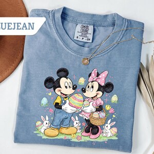 May include: Bluejean colored t-shirt featuring Mickey and Minnie Mouse holding Easter eggs. The graphic includes Easter eggs, bunnies, and flowers. A gold necklace is draped across the shirt. The text "BLUEJEAN" is in a white box in the upper left corner.