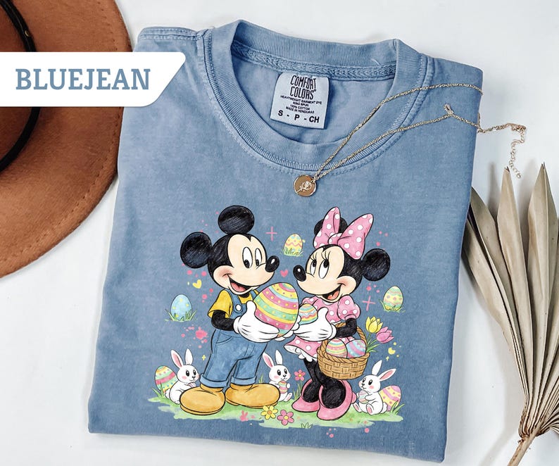 May include: Bluejean colored t-shirt featuring Mickey and Minnie Mouse holding Easter eggs. The graphic includes Easter eggs, bunnies, and flowers. A gold necklace is draped across the shirt. The text "BLUEJEAN" is in a white box in the upper left corner.