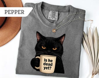 May include: A gray t-shirt featuring a black cat with yellow eyes holding a mug. The mug has the text "Is he dead yet?" printed on it. A silver necklace with a pendant is on the shirt.