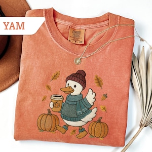 May include: A coral-colored Comfort Colors t-shirt featuring an embroidered duck wearing a hat, scarf, and boots, holding a coffee cup, and walking past pumpkins and autumn leaves. A gold necklace and a brown hat are also visible.