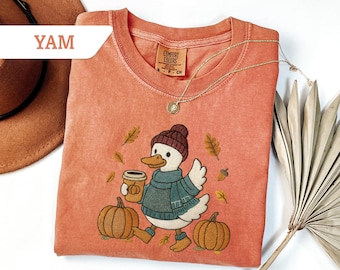 May include: A coral-colored Comfort Colors t-shirt featuring an embroidered duck wearing a hat, scarf, and boots, holding a coffee cup, and walking past pumpkins and autumn leaves. A gold necklace and a brown hat are also visible.