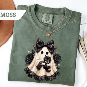 May include: A moss green short-sleeve t-shirt featuring a graphic of a ghost wearing a large leopard print bow and holding a black kitten. The ghost is surrounded by black roses and small bows. The shirt has a Comfort Colors brand label.
