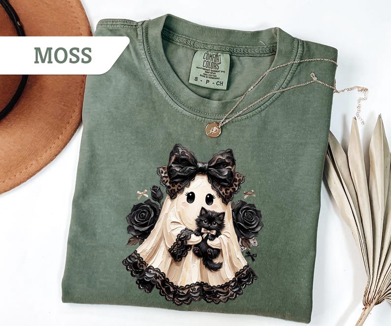 May include: A moss green short-sleeve t-shirt featuring a graphic of a ghost wearing a large leopard print bow and holding a black kitten. The ghost is surrounded by black roses and small bows. The shirt has a Comfort Colors brand label.