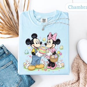 May include: Light blue t-shirt featuring Mickey and Minnie Mouse holding Easter eggs. The graphic includes bunnies, flowers, and the text "Chambray". The shirt is part of the Comfort Colors collection.