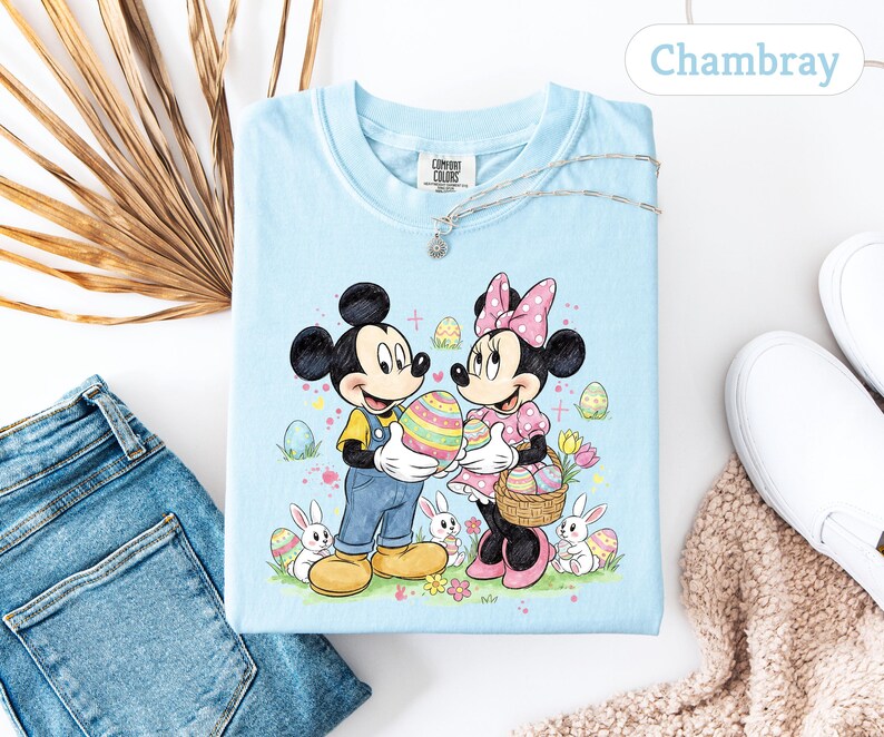 May include: Light blue t-shirt featuring Mickey and Minnie Mouse holding Easter eggs. The graphic includes bunnies, flowers, and the text "Chambray". The shirt is part of the Comfort Colors collection.