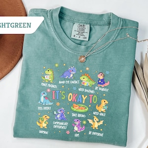 May include: Light green Comfort Colors t-shirt with the text "IT'S OKAY TO" and various dinosaur illustrations. The shirt also features the phrases "Make Mistakes", "Avoid Eye Contact", and "Be Yourself". A silver necklace is draped across the shirt.