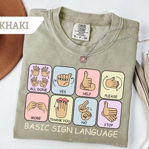 May include: Khaki-colored t-shirt with a "Basic Sign Language" design. The design features colorful squares with hand gestures and corresponding words like "All Done," "Yes," and "Help." A silver necklace with a pendant rests on the shirt.
