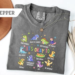 May include: Pepper-colored t-shirt with a dinosaur graphic and the text "It's Okay To." The graphic features colorful dinosaurs with phrases like "Make Mistakes" and "Be Yourself." A silver necklace with a pendant is draped on the shirt.