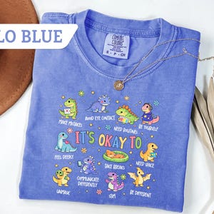 May include: A blue Comfort Colors t-shirt with the text "It's Okay To" and various dinosaur illustrations. The shirt also features the text "Flo Blue" in a white banner.