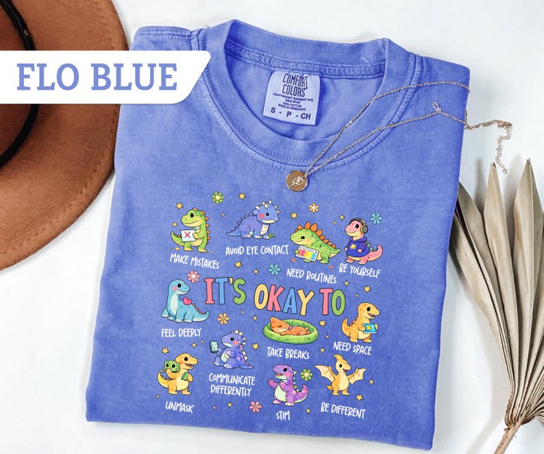 May include: A blue Comfort Colors t-shirt with the text "It's Okay To" and various dinosaur illustrations. The shirt also features the text "Flo Blue" in a white banner.