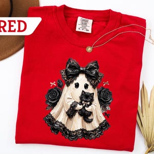 May include: A red crewneck t-shirt featuring a graphic of a ghost wearing a large black bow and holding a small black kitten. The ghost is surrounded by black roses and small bows. The shirt has a Comfort Colors label showing size S-CH.