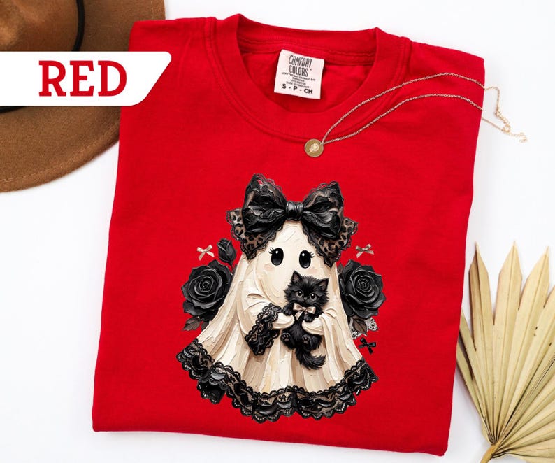 May include: A red crewneck t-shirt featuring a graphic of a ghost wearing a large black bow and holding a small black kitten. The ghost is surrounded by black roses and small bows. The shirt has a Comfort Colors label showing size S-CH.