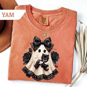 May include: A burnt orange short-sleeve t-shirt featuring a graphic of a ghost wearing a large black bow and holding a fluffy black kitten. The ghost is surrounded by black roses and lace trim. The shirt has a Comfort Colors brand label.