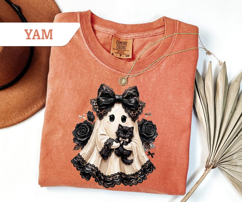 May include: A burnt orange short-sleeve t-shirt featuring a graphic of a ghost wearing a large black bow and holding a fluffy black kitten. The ghost is surrounded by black roses and lace trim. The shirt has a Comfort Colors brand label.