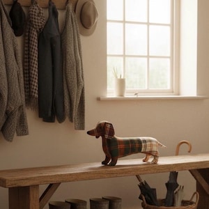 May include: A decorative dachshund dog figurine made of fabric with a plaid pattern in shades of brown, green, and cream. The dog is standing on a wooden bench. The background includes a coat rack, window, and a basket with umbrellas.