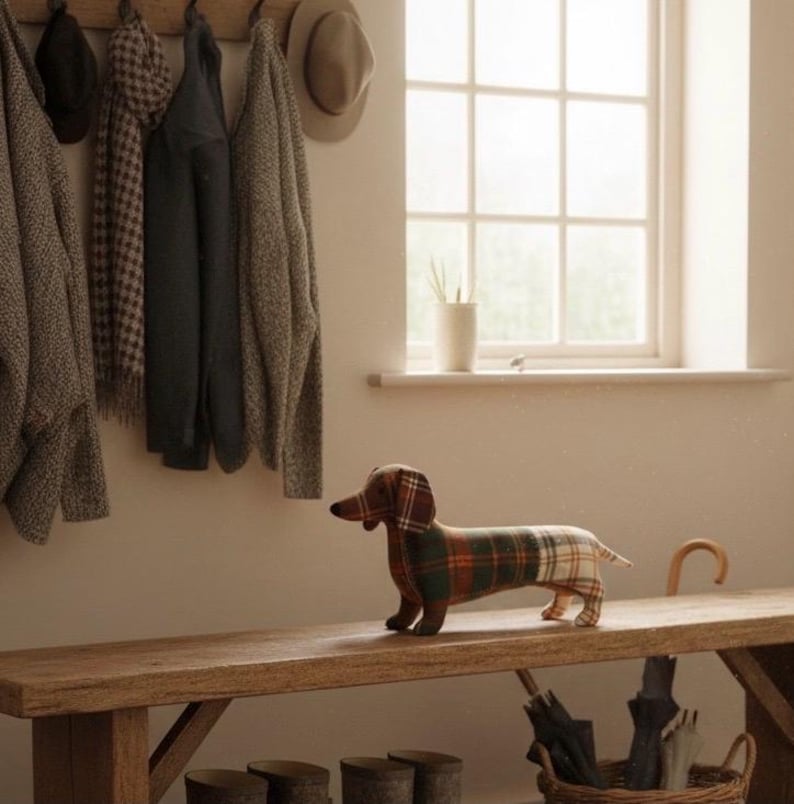 May include: A decorative dachshund dog figurine made of fabric with a plaid pattern in shades of brown, green, and cream. The dog is standing on a wooden bench. The background includes a coat rack, window, and a basket with umbrellas.