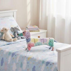 May include: A colorful dachshund-shaped pillow with tree and leaf patterns rests on a light blue bedspread. Other plush toys, including a sheep and an elephant, are also on the bed. The bed has a white headboard and is near a white nightstand.
