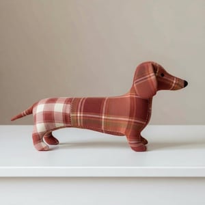 May include: A stuffed dachshund dog in a plaid pattern of red, white, and brown. The dog has a long body, short legs, and a black nose. The toy is made of fabric and sits on a white surface.