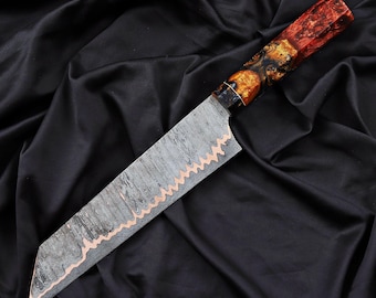 May include: A handcrafted chef's knife with a patterned steel blade and a copper-colored edge. The handle is a mix of red, orange, and brown tones. The knife is displayed on a black satin surface.