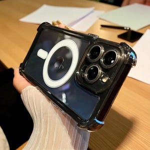 May include: Clear phone case with a black border and a white circular design on the back. The case has a built-in camera protector and is designed to fit a smartphone. The case is held in a person's hand.