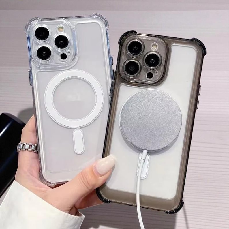 May include: Two clear phone cases with protective bumpers. One case has a white magnetic charging ring and a white charging port. The other case has a silver magnetic charging ring and a white charging port.