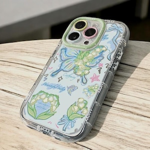 May include: A clear phone case with a blue and white butterfly design. The case has a green and pink ring around the camera lens. The case also has a white background with a variety of other designs, including flowers, stars, and the words "Gathering" and "Beautiful Tulip".