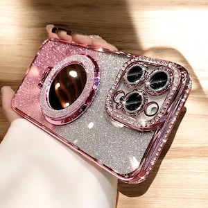 May include: Pink and silver glitter phone case with a ring holder and a camera lens protector. The case has a sparkly, shiny appearance with a pink border and is embellished with rhinestones.