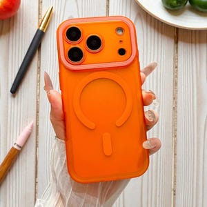 Peut inclure: Coque de téléphone orange vif avec un design d'anneau magnétique. L'étui a un bord surélevé autour des lentilles de la caméra. L'étui est tenu dans une main avec de longs ongles manucurés. Un stylo et un marqueur rose sont en arrière-plan.