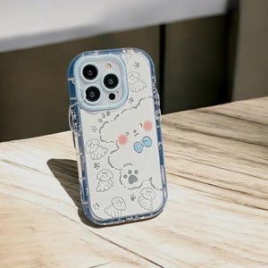 May include: A clear phone case with a white background and a cute cartoon dog design. The case has a blue trim and a cutout for the camera lens.